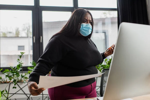 Afroamericana más tamaño mujer de negocios en máscara médica celebración de teléfono inteligente y papel mientras mira el monitor de la computadora - foto de stock