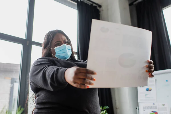 Africano americano más tamaño mujer de negocios en máscara médica celebración de papel con gráficos y gráficos - foto de stock