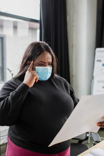 Africano americano más tamaño mujer de negocios en médico máscara celebración de papel en la oficina - foto de stock