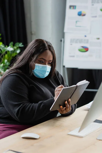 Afroamericano plus size donna d'affari in maschera medica in possesso di notebook vicino al computer — Foto stock