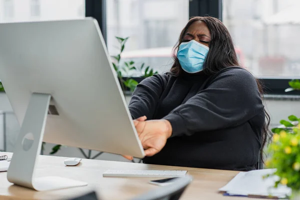 Stanco africano americano plus size donna d'affari in maschera medica che si estende vicino al monitor del computer — Foto stock
