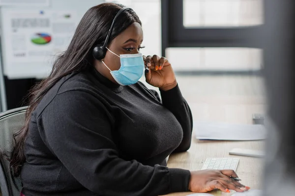 Afroamericano más operador de tamaño en auriculares y máscara médica hablando en la oficina - foto de stock