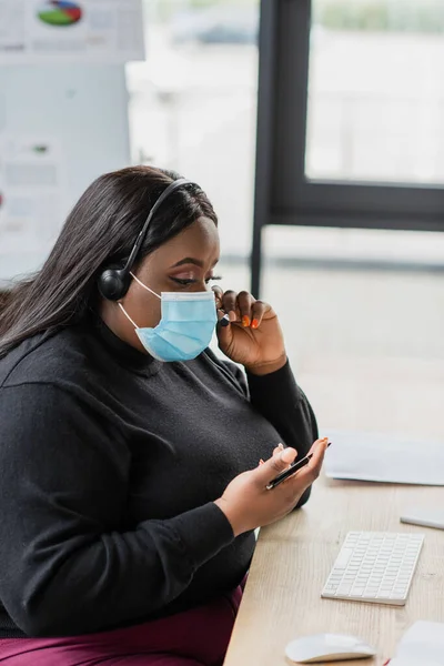 Africano americano más operador de tamaño en auriculares y máscara médica hablando en micrófono - foto de stock