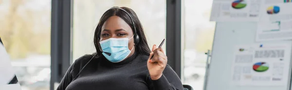 Афроамериканський оператор плюс розмір в гарнітурі та медичній масці тримає ручку в сучасному офісі, банер — стокове фото