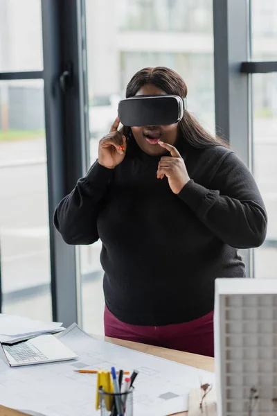 Stupito africano americano plus size donna in auricolare vr vicino modello di casa in cartone e cianografia sulla scrivania — Foto stock