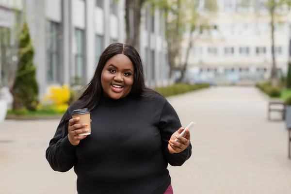 Feliz afroamericano más tamaño mujer sosteniendo café para ir y teléfono inteligente fuera - foto de stock