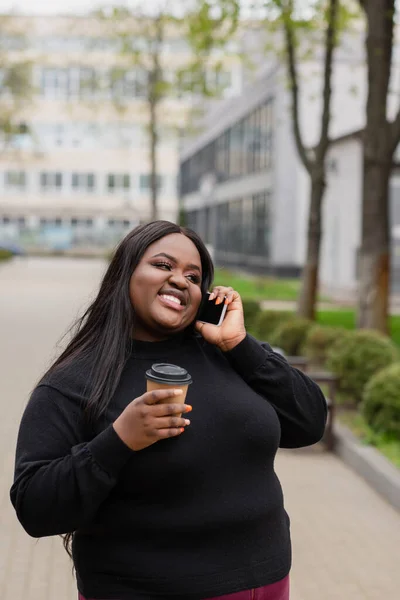 Alegre afroamericano más tamaño mujer sosteniendo café para ir y hablar en el teléfono inteligente fuera - foto de stock