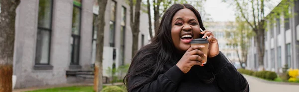 Alegre afroamericano más tamaño mujer sosteniendo café para ir y hablar en el teléfono inteligente fuera, pancarta - foto de stock