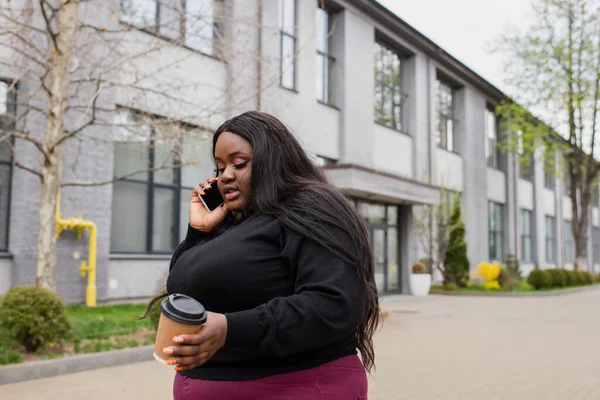 Cheerful african american plus size woman holding notebook and speaking on smartphone outside — Stock Photo