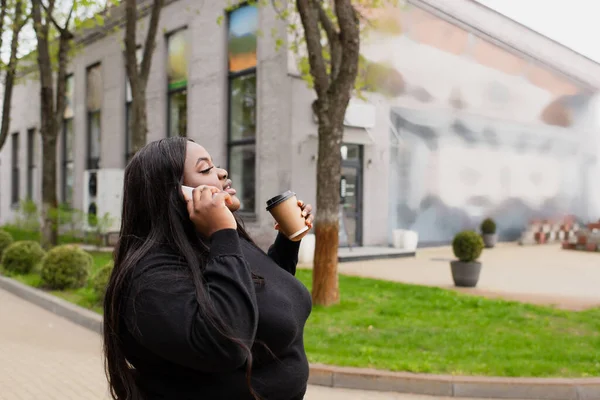 Vista lateral de afroamericano más tamaño mujer sosteniendo taza de papel y hablando en el teléfono inteligente fuera - foto de stock