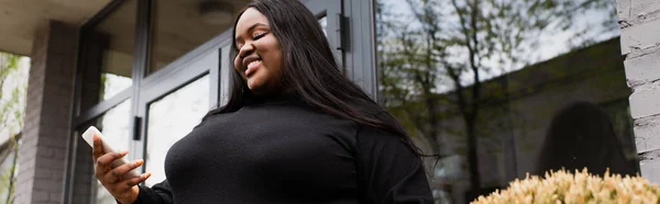 Low angle view of cheerful african american plus size woman holding coffee to go and looking at smartphone outside, banner — Stock Photo