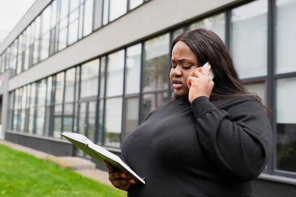 Afro-américaine plus taille femme tenant un cahier et parlant sur smartphone à l'extérieur — Photo de stock