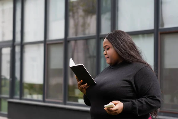African american plus size woman holding notebook and smartphone outside — Stock Photo