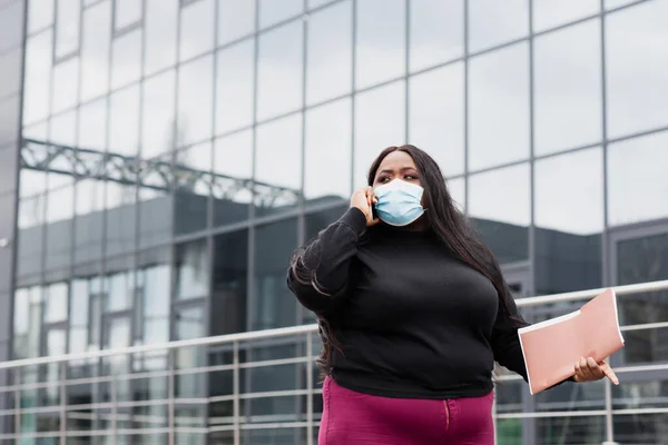 Afro americano plus size donna in maschera medica tenendo cartella e parlando su smartphone al di fuori — Foto stock