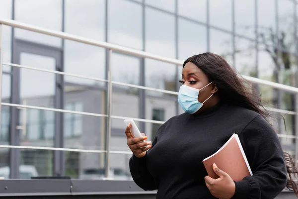 Africano americano más tamaño mujer de negocios en máscara médica celebración de la carpeta y el teléfono inteligente fuera - foto de stock