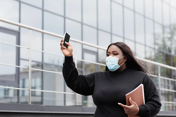 Africano americano más tamaño mujer en médico máscara celebración carpeta y teléfono inteligente con pantalla en blanco fuera - foto de stock