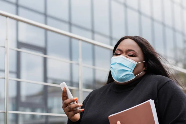 Africano americano más tamaño mujer en médico máscara celebración carpeta y teléfono celular fuera - foto de stock