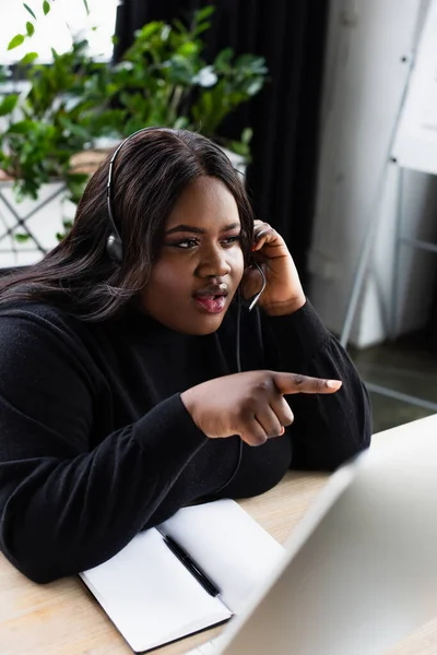 Afrikanischer Plus-Size-Bediener im Headset mit Mikrofon, das mit dem Finger auf verschwommenen Computermonitor zeigt — Stockfoto