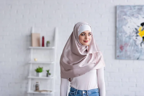 Junge muslimische Frau im Hidschab lächelt, während sie zu Hause wegschaut — Stockfoto