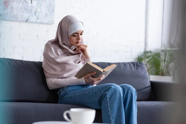 Mujer musulmana bastante leer libro en el sofá cerca de taza borrosa de té - foto de stock