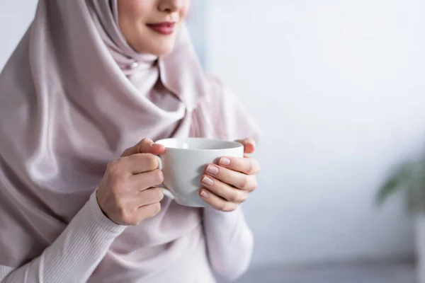 Vista cortada de mulher muçulmana desfocada em hijab segurando xícara de chá em casa — Fotografia de Stock