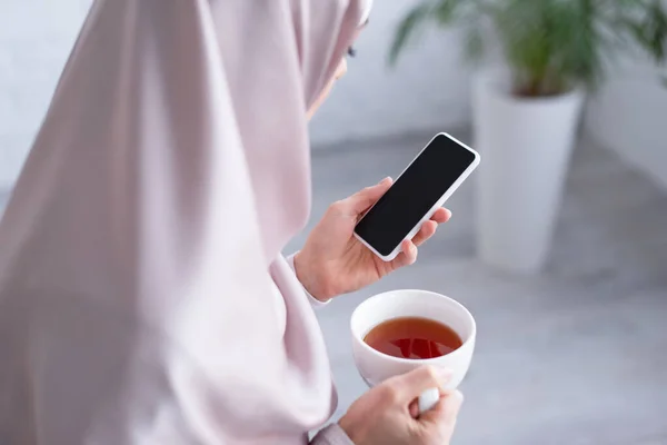 Femme musulmane tenant tasse de thé et téléphone portable avec écran blanc à la maison — Photo de stock