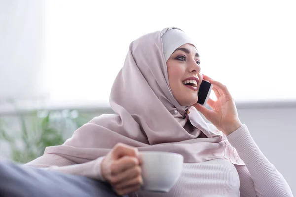 Cheerful muslim woman with cup of tea talking on smartphone at home — Stock Photo