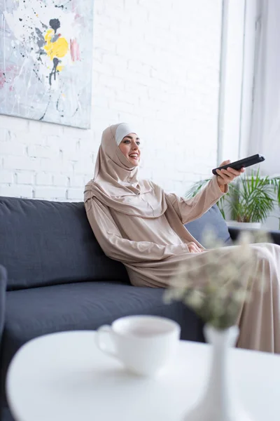 Donna musulmana sorridente che guarda la tv sul divano vicino alla tazza di tè offuscata sul tavolino — Foto stock