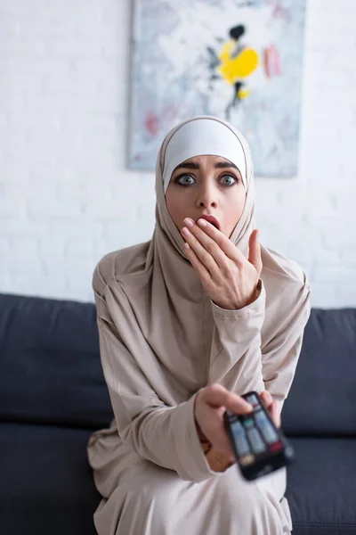 Schockierte Muslimin bedeckt Mund mit der Hand, während sie zu Hause fernsieht — Stockfoto