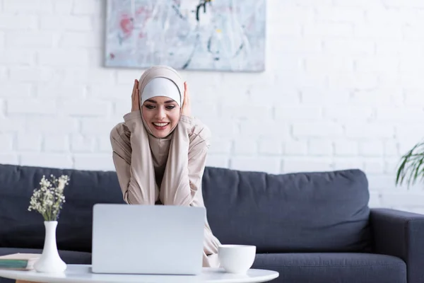 Mulher muçulmana sorrindo enquanto olha para laptop em casa — Fotografia de Stock