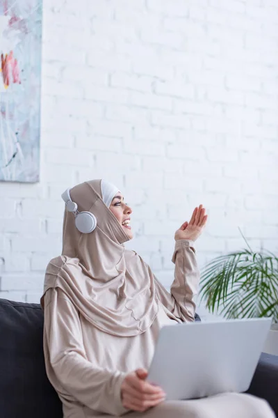 Mulher muçulmana alegre em fones de ouvido gesticulando perto de laptop em casa — Fotografia de Stock