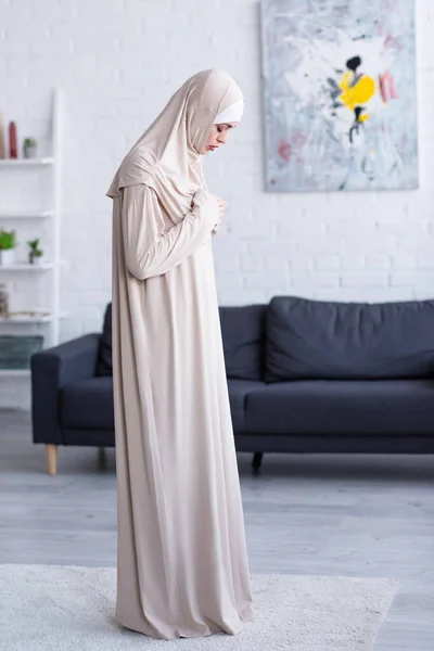 Muslim woman praying with hands on chest while standing in living room — Stock Photo