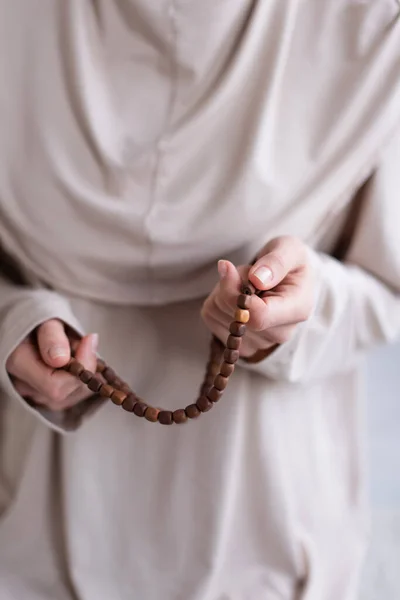 Selektiver Fokus des Rosenkranzes in den Händen einer verschwommenen muslimischen Frau, die zu Hause betet, abgeschnittene Ansicht — Stockfoto