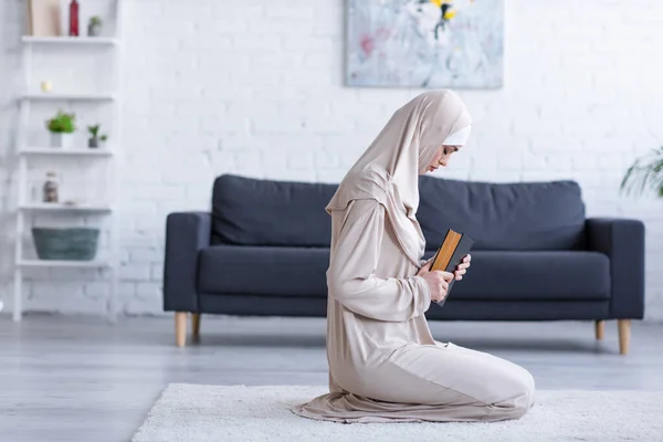 Vista laterale della donna musulmana che prega con il Corano sul pavimento a casa — Foto stock