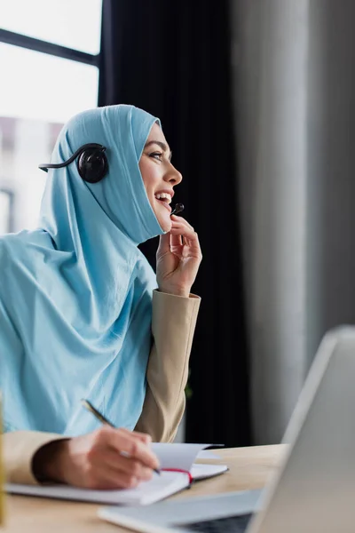 Alegre árabe mujer en hijab y auriculares trabajando en centro de llamadas cerca borrosa portátil - foto de stock