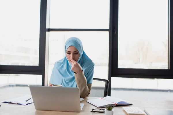 Pensativa mujer de negocios musulmana que trabaja cerca de la computadora portátil contra la ventana en la oficina - foto de stock