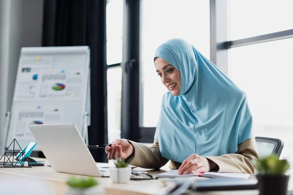 Lächelnde muslimische Geschäftsfrau zeigt auf Laptop, während sie im Büro arbeitet, verschwommener Vordergrund — Stockfoto