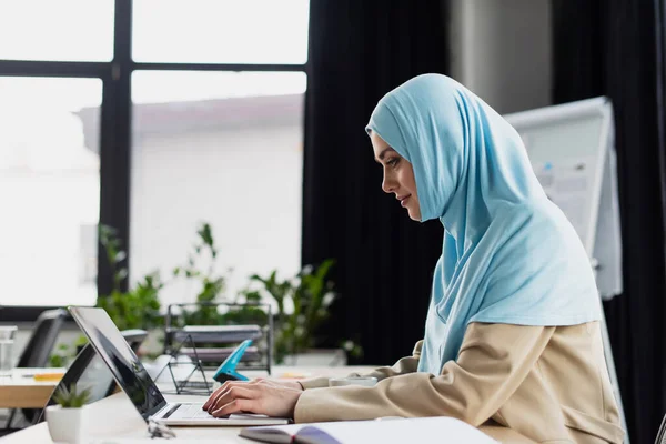 Vista lateral de la joven mujer de negocios árabe en hijab escribiendo en el ordenador portátil en la oficina - foto de stock