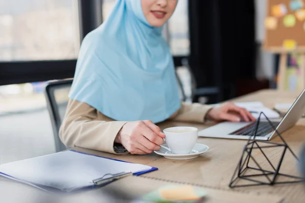 Vue recadrée d'une femme d'affaires musulmane travaillant près d'un ordinateur portable et d'une tasse de café au premier plan flou — Photo de stock