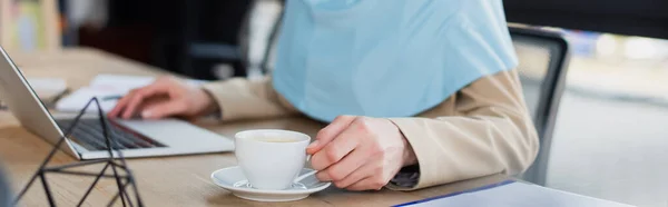 Обрізаний вид мусульманської бізнес-леді біля ноутбука та чашки кави в офісі, банер — Stock Photo