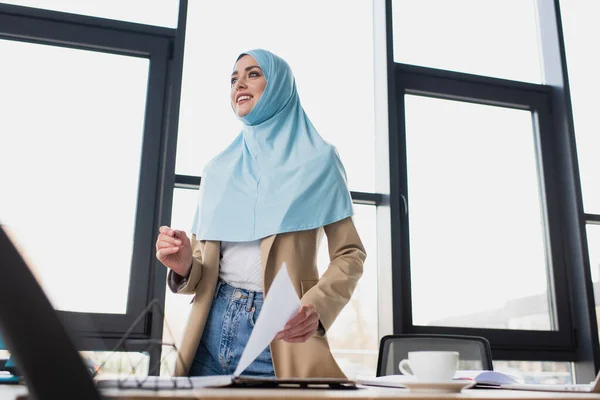 Mujer de negocios musulmana sonriente sosteniendo papel mientras está de pie contra la ventana en la oficina - foto de stock
