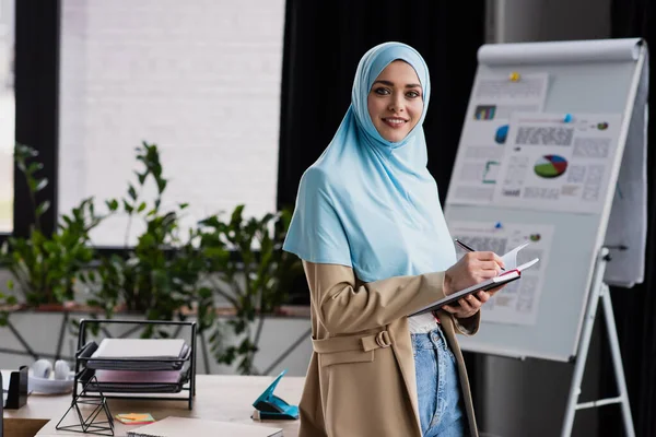 Feliz musulmana mujer de negocios escribiendo en cuaderno cerca de rotafolio sobre fondo borroso - foto de stock