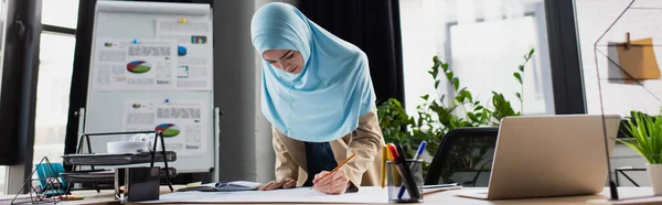 Dibujo de ingeniero musulmán con lápiz cerca del ordenador portátil e infografías en el fondo, pancarta - foto de stock