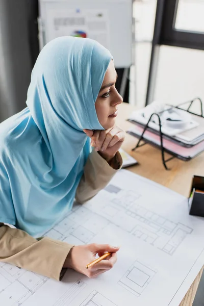 Pensativo ingeniero musulmán sosteniendo lápiz cerca de plano en la oficina - foto de stock