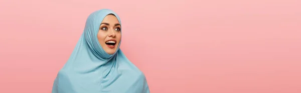 Femme arabe étonnée en hijab bleu regardant loin isolé sur rose, bannière — Photo de stock