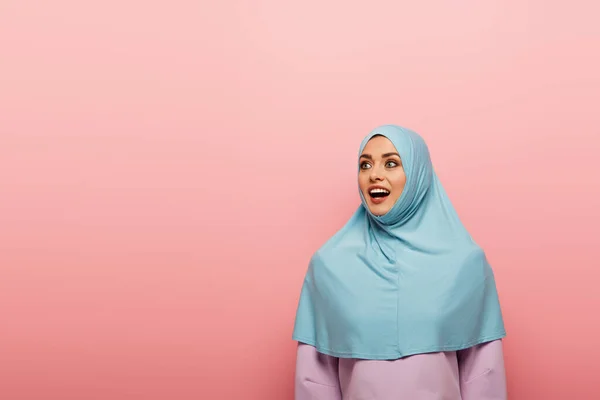 Emocionada mujer musulmana en hijab azul mirando hacia otro lado aislado en rosa - foto de stock
