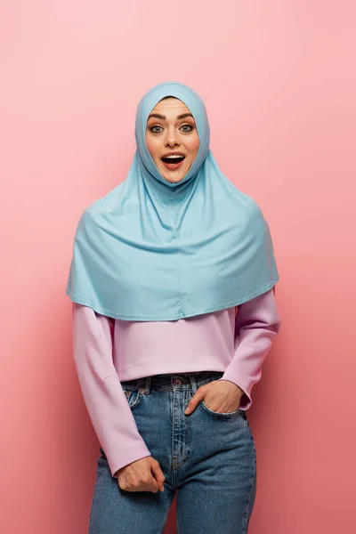 Amazed muslim woman in jeans standing with hand in pocket on pink background — Stock Photo