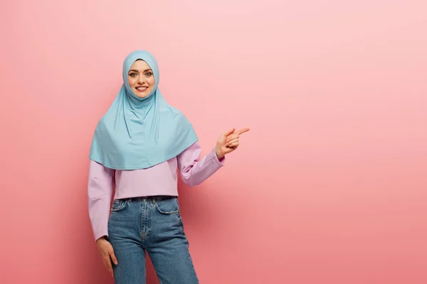 Mujer árabe alegre en jeans y hijab señalando a un lado con el dedo sobre fondo rosa - foto de stock