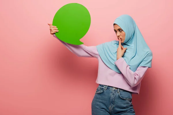 Nachdenkliche muslimische Frau, die Lippen berührt, während sie grüne Gedankenblase auf rosa Hintergrund betrachtet — Stockfoto