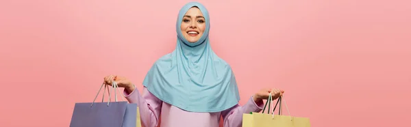 Довольная мусульманка с сумками для покупок, глядя на камеру, изолированную на розовый, баннер — стоковое фото